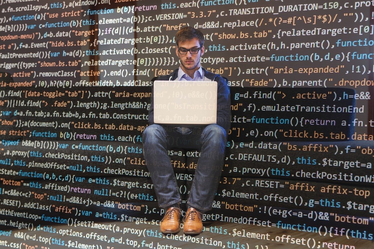 Un joven programador con un ordenador portátil sobre sus rodillas sobre un fondo de código JavaScript